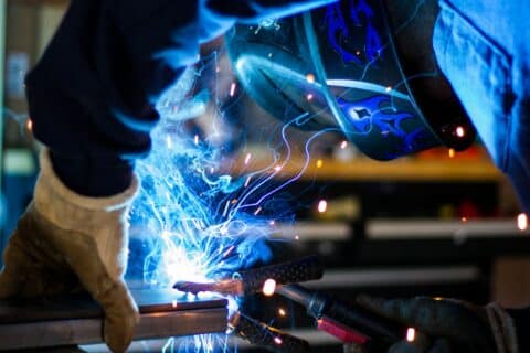 Welder working on a metal fabrication project with welding sparks visible at the work area.